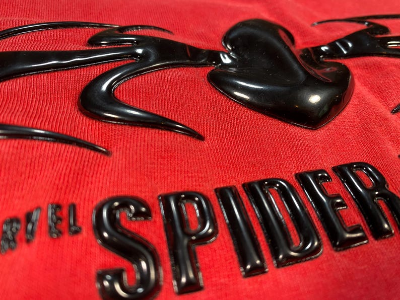 Op de afbeelding: Close-up van een rode stof met een glanzend zwart ontwerp. Het ontwerp bevat het woord "SPIDER" in vette letters en een gestileerde hartvorm. De stof heeft een getextureerd uiterlijk.
