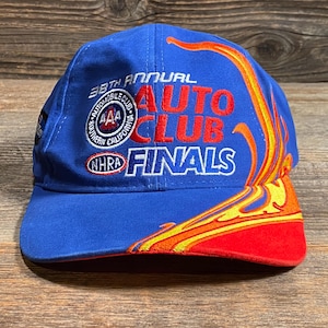 Puede incluir: Gorra de b&eacute;isbol azul con el texto "38th Annual Auto Club Finals" en rojo y blanco. La gorra presenta un dise&ntilde;o de llama en amarillo, naranja y rojo. La visera es roja.
