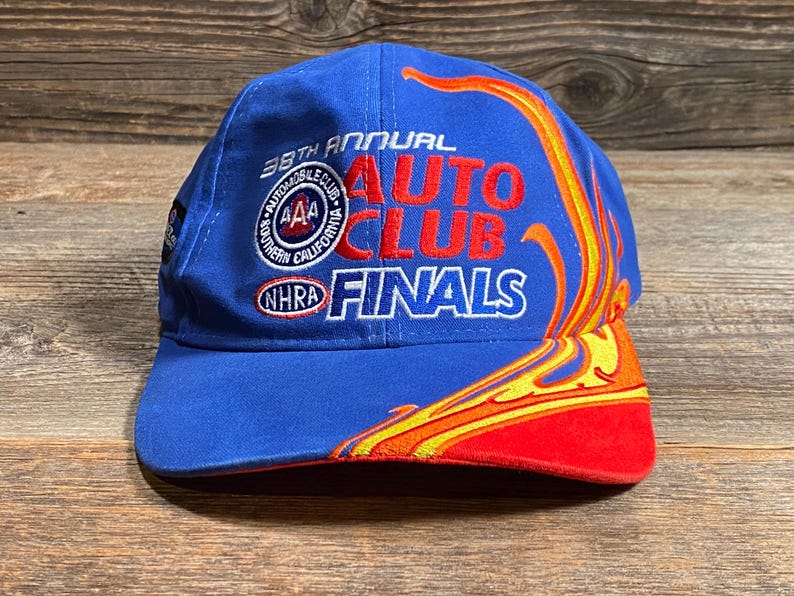Puede incluir: Gorra de b&eacute;isbol azul con el texto "38th Annual Auto Club Finals" en rojo y blanco. La gorra presenta un dise&ntilde;o de llama en amarillo, naranja y rojo. La visera es roja.