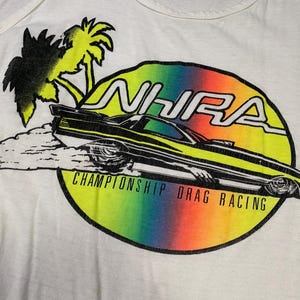 Pu&ograve; includere: Canotta bianca con una grafica colorata di un'auto da drag racing. Il design include il testo "NHRA CHAMPIONSHIP DRAG RACING", un gradiente arcobaleno, palme e un'auto da corsa stilizzata.