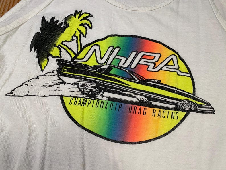 Pu&ograve; includere: Canotta bianca con una grafica colorata di un'auto da drag racing. Il design include il testo "NHRA CHAMPIONSHIP DRAG RACING", un gradiente arcobaleno, palme e un'auto da corsa stilizzata.