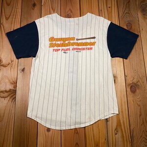 Peut inclure: Un maillot de baseball &agrave; manches courtes avec un corps couleur cr&egrave;me orn&eacute; de fines rayures verticales noires et des manches bleu marine unies. L'avant pr&eacute;sente le texte orange "Gunner Steinbrenner" avec une batte et "TOP FUEL DRAGSTER".
