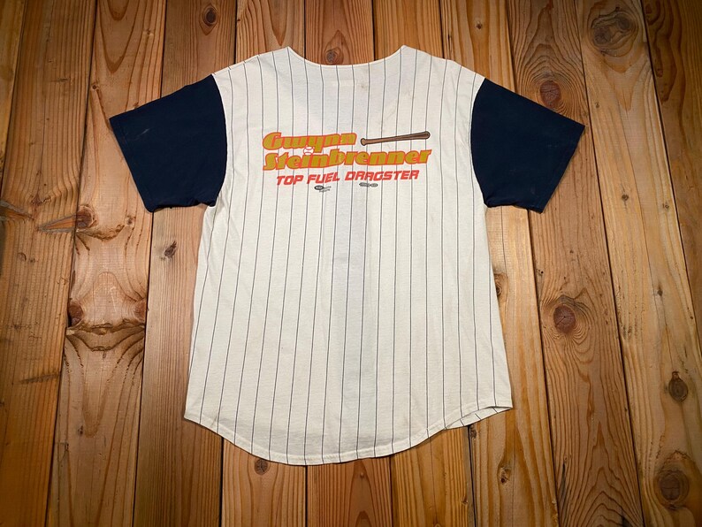 Peut inclure: Un maillot de baseball &agrave; manches courtes avec un corps couleur cr&egrave;me orn&eacute; de fines rayures verticales noires et des manches bleu marine unies. L'avant pr&eacute;sente le texte orange "Gunner Steinbrenner" avec une batte et "TOP FUEL DRAGSTER".