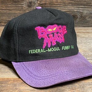May include: Black cap with a purple brim. The cap features the embroidered text "Boogie Man" in pink and the text "FEDERAL-MOGUL FUNNY CAR" in green. The cap has a purple button on top.