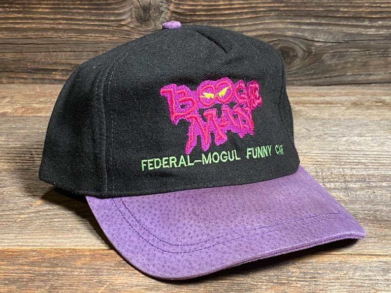 May include: Black cap with a purple brim. The cap features the embroidered text "Boogie Man" in pink and the text "FEDERAL-MOGUL FUNNY CAR" in green. The cap has a purple button on top.