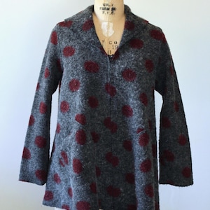 May include: A charcoal grey jacket with a textured, felted appearance featuring a pattern of dark red polka dots. The jacket has long sleeves, a collared neckline, and a central zip closure that extends from the hem to the collar.