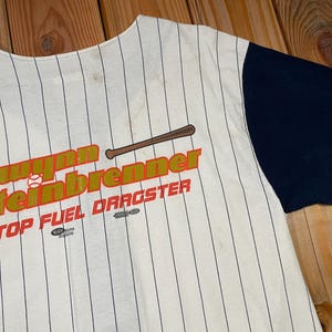 Peut inclure: Un maillot de baseball couleur cr&egrave;me avec de fines rayures noires et des manches bleu marine unies. L'avant pr&eacute;sente un motif avec une batte de baseball marron, une petite balle de baseball blanche et le texte Gwynn Steinbrenner TOP FUEL DRAGSTER.
