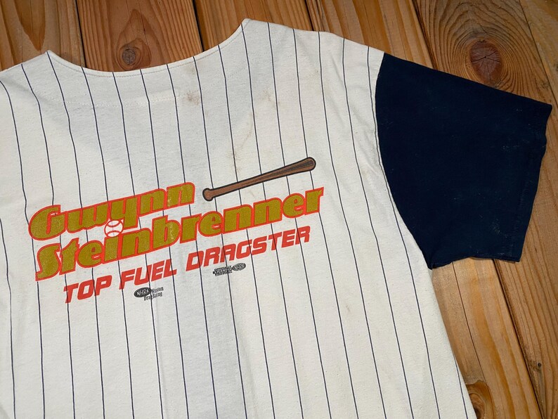 Peut inclure: Un maillot de baseball couleur cr&egrave;me avec de fines rayures noires et des manches bleu marine unies. L'avant pr&eacute;sente un motif avec une batte de baseball marron, une petite balle de baseball blanche et le texte Gwynn Steinbrenner TOP FUEL DRAGSTER.
