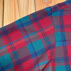 May include: Close-up of a plaid shirt sleeve. The fabric features a pattern of red, teal, blue, and brown squares and lines. The sleeve is sewn at the shoulder. The background is a light brown wooden surface.