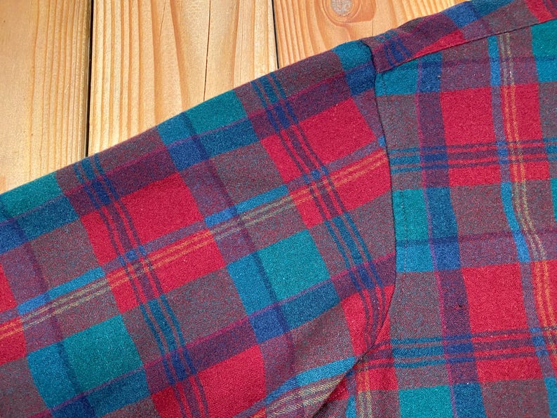 May include: Close-up of a plaid shirt sleeve. The fabric features a pattern of red, teal, blue, and brown squares and lines. The sleeve is sewn at the shoulder. The background is a light brown wooden surface.