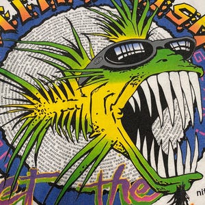 Puede incluir: Camiseta blanca con un dise&ntilde;o de pez de dibujos animados. El pez, con gafas de sol, tiene dientes afilados y es verde y amarillo. El dise&ntilde;o incluye el texto "ULTIMATE GEAR" y "GET THE" en un borde circular.