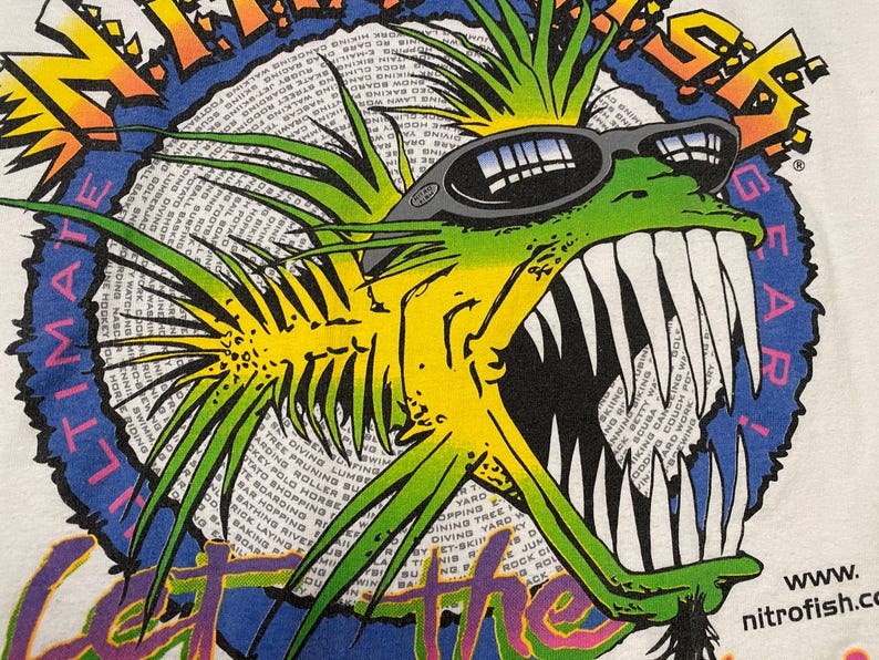 Puede incluir: Camiseta blanca con un dise&ntilde;o de pez de dibujos animados. El pez, con gafas de sol, tiene dientes afilados y es verde y amarillo. El dise&ntilde;o incluye el texto "ULTIMATE GEAR" y "GET THE" en un borde circular.