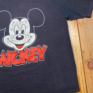 Puede incluir: Una camiseta negra de manga corta con un gr&aacute;fico grande y desgastado en blanco del rostro de Mickey Mouse. Debajo del personaje, la palabra MICKEY est&aacute; impresa en letras may&uacute;sculas rojas desgastadas con un borde blanco.