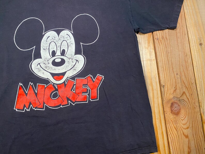 Puede incluir: Una camiseta negra de manga corta con un gr&aacute;fico grande y desgastado en blanco del rostro de Mickey Mouse. Debajo del personaje, la palabra MICKEY est&aacute; impresa en letras may&uacute;sculas rojas desgastadas con un borde blanco.