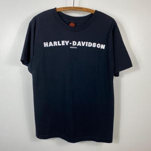 May include: Black Harley-Davidson t-shirt with white lettering. The brand name is prominently displayed across the chest. The shirt is hanging on a wooden hanger against a plain background. The shirt has a crew neck and short sleeves.
