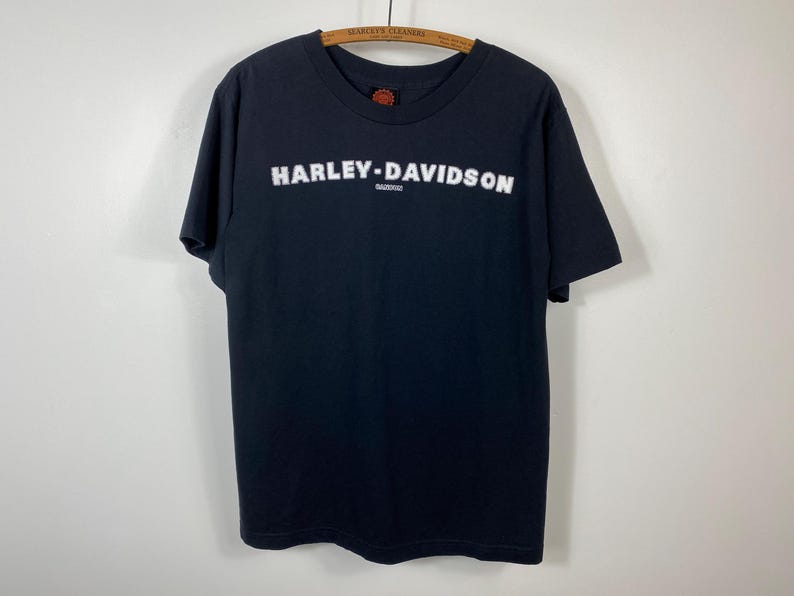 May include: Black Harley-Davidson t-shirt with white lettering. The brand name is prominently displayed across the chest. The shirt is hanging on a wooden hanger against a plain background. The shirt has a crew neck and short sleeves.