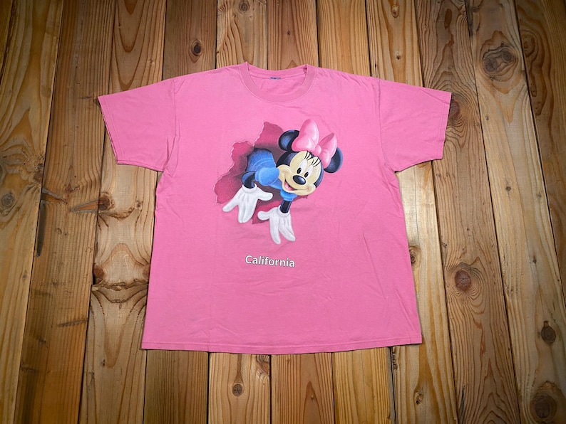 May include: A bright pink short-sleeved t-shirt featuring a graphic of Minnie Mouse emerging from a dark red cutout shape. Minnie Mouse wears a pink bow and white gloves. The word California is printed in white text below the graphic.