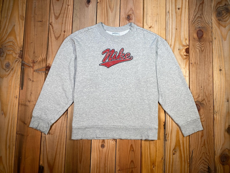 Puede incluir: Una sudadera gris jaspeada con el logo de Nike en rojo. La parte superior de manga larga y cuello redondo est&aacute; hecha de un material suave. La sudadera se muestra sobre una superficie de madera.