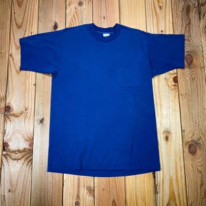 Könnte beinhalten: Ein einfarbiges blaues T-Shirt mit einer Brusttasche. Das kurzärmelige Shirt besteht aus einem weichen Material und wird auf einer Holzoberfläche präsentiert. Das Shirt ist ein klassischer, lässiger Stil.
