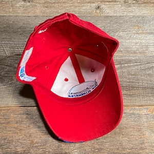 Op de afbeelding: Een rode honkbalpet gezien vanaf de binnenkant, voorzien van een witte voering met rode steunbanden, een centrale metalen knop en een wit stoffen label met blauwe tekst. Een cirkelvormig logo-embleem is zichtbaar aan de binnenzijde.