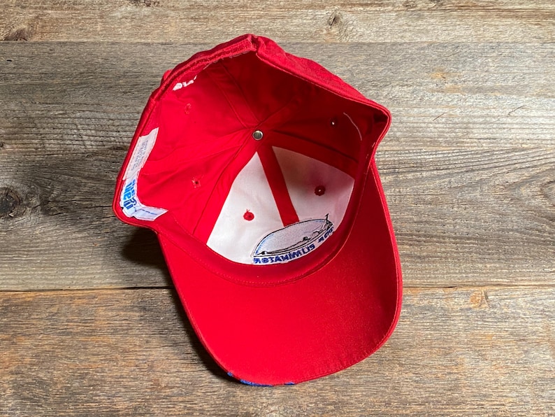 Op de afbeelding: Een rode honkbalpet gezien vanaf de binnenkant, voorzien van een witte voering met rode steunbanden, een centrale metalen knop en een wit stoffen label met blauwe tekst. Een cirkelvormig logo-embleem is zichtbaar aan de binnenzijde.