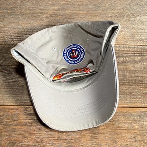 May include: A pale grey baseball cap with a curved peak and a circular patch. The patch reads "AUTOMOBILE CLUB" and "SOUTHERN CALIFORNIA" around a central "AAA" logo. The cap has a white trim on the peak.