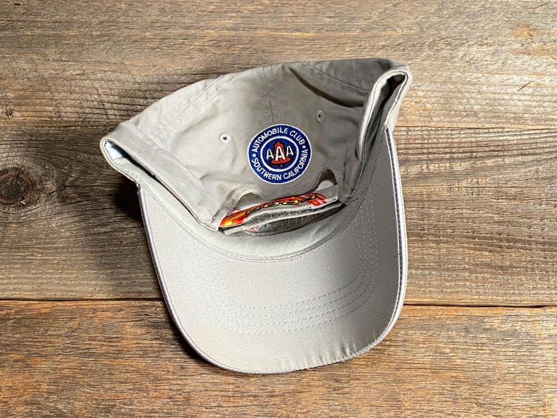 May include: A pale grey baseball cap with a curved peak and a circular patch. The patch reads "AUTOMOBILE CLUB" and "SOUTHERN CALIFORNIA" around a central "AAA" logo. The cap has a white trim on the peak.