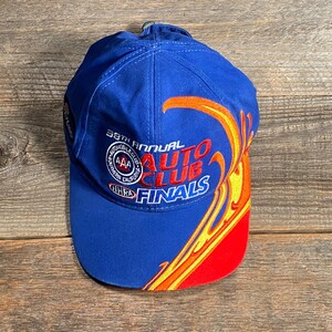 Puede incluir: Gorra de b&eacute;isbol azul con el texto "38th Annual Auto Club Finals" en rojo y blanco. La gorra presenta un dise&ntilde;o de llama en amarillo, naranja y rojo. El sombrero est&aacute; hecho de una tela duradera.