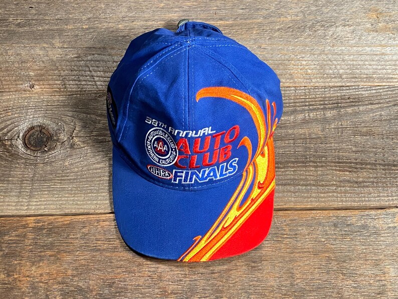 Puede incluir: Gorra de b&eacute;isbol azul con el texto "38th Annual Auto Club Finals" en rojo y blanco. La gorra presenta un dise&ntilde;o de llama en amarillo, naranja y rojo. El sombrero est&aacute; hecho de una tela duradera.