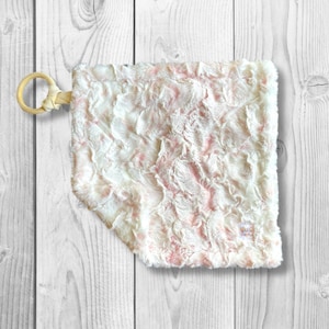 May include: A soft, white and pink baby blanket with a knotted wooden ring attached. The blanket has a fluffy, textured surface and a small tag with the text "Bubba Love" on it.