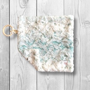 May include: A soft, white and teal plush baby blanket with a wooden ring attached. The blanket has a crinkled texture.