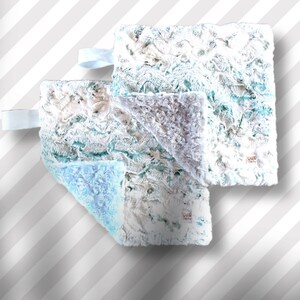 May include: Three plush baby blankets in shades of white, blue, and teal. The blankets have a textured, rose-like pattern and a satin ribbon loop. One blanket is folded to reveal a light blue underside. The blankets are soft and fluffy.