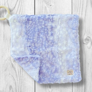 May include: A soft, lavender and white plush baby blanket with a wooden ring attached. The blanket has a subtle, speckled pattern and is perfect for cuddling.