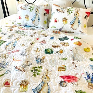May include: White quilted bedding set featuring a whimsical Peter Rabbit design. The set includes two pillowcases and a quilt adorned with illustrations of rabbits, vegetables, and kitchen tools. The quilt is patterned with a repeating circular design.