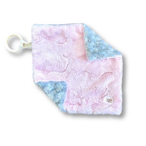 May include: A soft, pink and blue baby blanket with a wooden ring attached. The blanket has a textured, wavy pattern and a small tag that says "Hello Little One".
