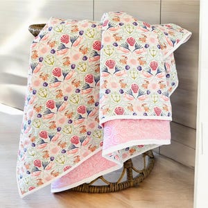 May include: Two decorative beach-themed blankets with a seashell, anchor, and crab pattern in pink, blue, and green. The blankets are folded over a wicker chair. The bottom blanket has a pink border with a swirl design.