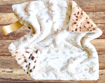 May include: A white and brown faux fur baby blanket with a brown and white spotted pattern. The blanket has a loop handle and is folded in half.