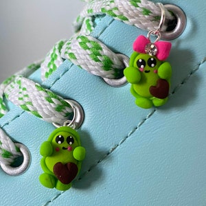 Könnte beinhalten: Zwei grüne Avocado-förmige Charms mit braunen Herzen und lächelnden Gesichtern. Ein Charm hat eine rosa Schleife und einen Strassstein. Die Charms sind an Schnürsenkeln an einem hellblauen Schuh befestigt.