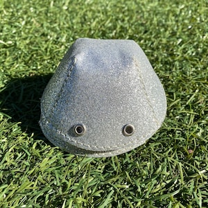 Puede incluir: Una cúpula de purpurina plateada con dos pequeños ojales plateados. La cúpula está sentada sobre un lecho de hierba verde.