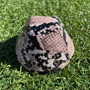 Puede incluir: Un sombrero de color rosa y negro con estampado de piel de serpiente y dos ojales plateados.
