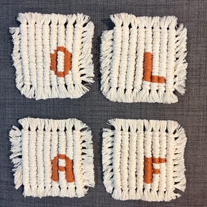 May include: A set of four square macrame coasters made from off-white cotton cord with fringed edges. Each coaster features a single large letter in a burnt orange colour, spelling out the word O-L-A-F across the four individual pieces.
