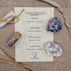 May include: A set of crystals and a bottle of lavender for spiritual practices. The set includes amethyst, quartz geode, selenite wand, and a bottle of lavender. The crystals are arranged on a piece of paper with the text "Little Box of Spirituality" and "Crystal Gift Set".
