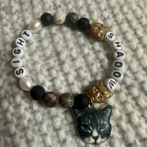 Op de afbeelding: Een zwarte, witte en groene kralen armband met een kattenbedel. De armband heeft de woorden "Sight" en "Shadow" erop.