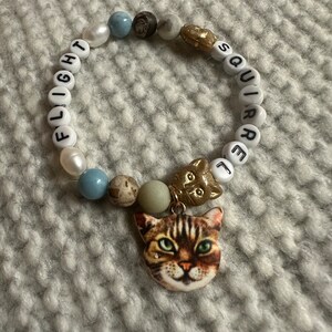 Op de afbeelding: Een witte kralen armband met de woorden "FLIGHT SQUIRREL" gespeld in zwarte letters. De armband heeft een bedel van een kattenkop met bruin en wit bont.