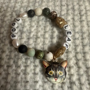 Op de afbeelding: Een kralenarmband met de naam "LEA" en "POOL" gespeld in zwarte letters op witte kralen. De armband heeft een verscheidenheid aan andere kralen, waaronder zwarte, witte, groene en bruine. Een kattenhanger hangt aan de armband.