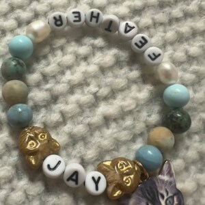 Op de afbeelding: Een witte kralen armband met het woord "FATHER" gespeld in zwarte letters. De armband heeft ook blauwe en bruine kralen, en twee gouden katten bedels met het woord "JAY" gespeld in witte letters.