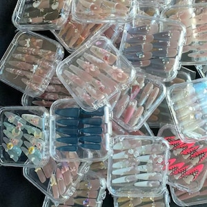 May include: An assortment of artificial nails in clear plastic cases. The nails vary in color, including pink, nude, and blue, and feature designs such as floral patterns, glitter, and rhinestones. Some nails are solid colors, while others have decorative elements.