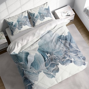 Pode incluir: Uma capa de edredom branca com um design abstrato de montanha azul e branco. A capa de edredom apresenta uma cordilheira estilizada com uma silhueta de veado. O design lembra uma pintura em aquarela.