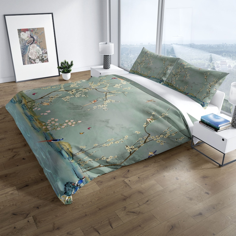 Puede incluir: Una funda n&oacute;rdica azul y blanca con un estampado floral que presenta un &aacute;rbol con flores blancas y p&aacute;jaros. La funda n&oacute;rdica est&aacute; en una cama con dos fundas de almohada a juego.