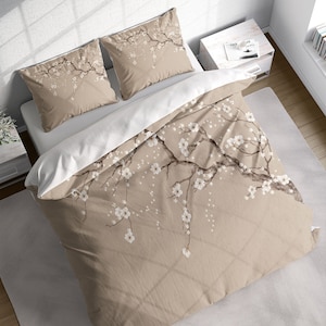Può includere: Un set di biancheria da letto floreale beige con un copripiumino e due federe. Il design presenta un'illustrazione dettagliata di un ramo di albero in fiore con fiori bianchi. Il copripiumino ha un lato inferiore bianco.